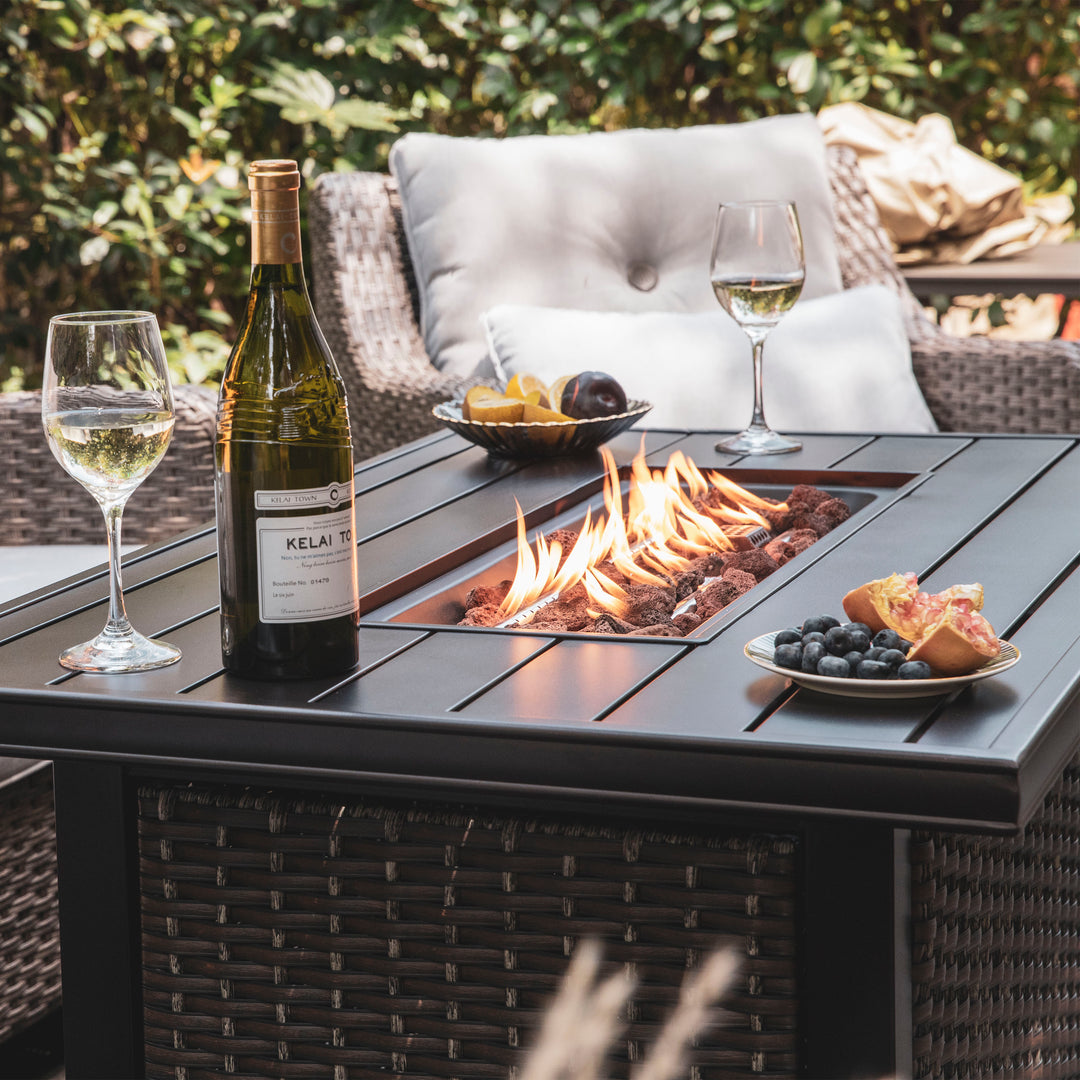 A wine bottle and a glass on a table with an integrated fire pit, alongside a plate of fruit and a cozy outdoor seating area in the background.